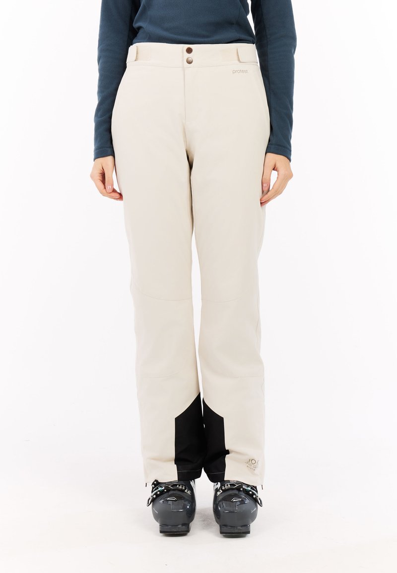 Beige skibroek met zwarte accenten, gemaakt van een zacht, flexibel materiaal. Voorzien van een knoopsluiting en een logo nabij de zoom. Soepele textuur.