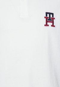 Vit polotröja med en texturerad tygtyp. Har en krage med tre knappar och en liten broderad logotyp i marinblått och röd på vänster bröst.