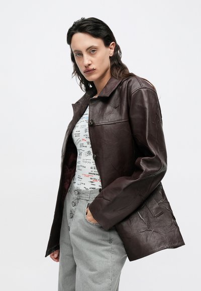 Bruine leren jas met een kraag en knoopsluiting aan de voorkant. De drager combineert het met een overhemd met patroon en grijze broek. Heeft een gladde textuur.