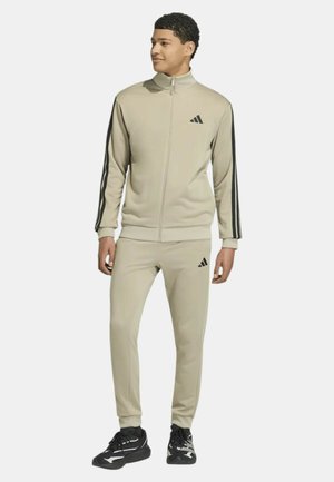 Giovane in piedi, indossa una giacca sportiva beige con cerniera integrale e pantaloni con strisce nere e logo, abbinati a scarpe sportive nere.