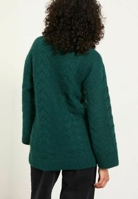 Person mit lockigem Haar, der einen dunkelgrünen strukturierten Strickpullover und schwarze Hosen trägt, mit dem Rücken zu einem schlichten Hintergrund gewandt.