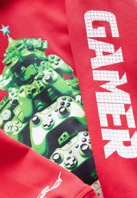 Sweatshirt rouge avec un graphique de manettes de jeu vertes, texte "GAMER" en blanc, avec une étoile en accent au-dessus. Texture de tissu douce.