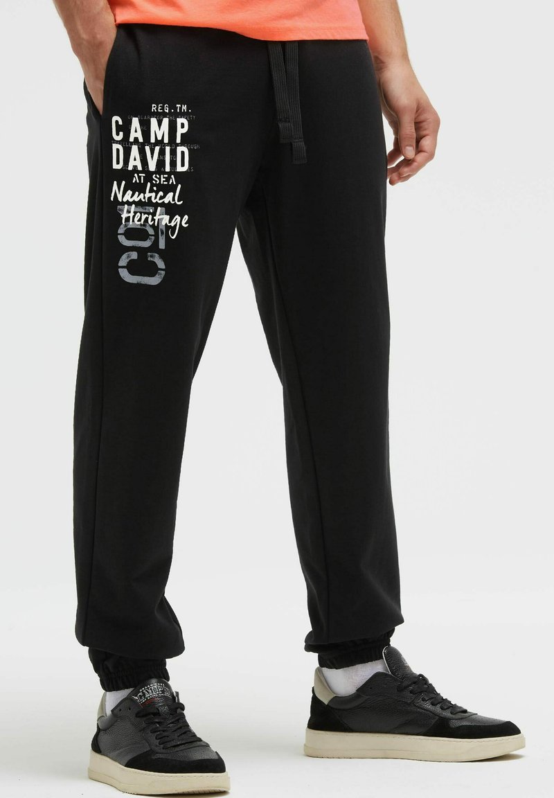 Schwarze Jogginghose mit elastischen Bündchen und verstellbarem Kordelzug, mit weißem Aufdruck und Grafikdesign am linken Bein.