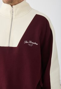 Pullover borgogna e crema con zip e colletto alto. Presenta un design triangolare a contrasto e testo ricamato sul fronte. Materiale in cotone.