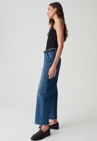 Zwarte tanktop gecombineerd met wijde blauwe denim broek. De denim heeft een gladde textuur met minimale details. Zwarte loafers maken de outfit compleet.