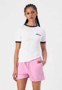 Hvit T-skjorte med kort erm og marineblå detaljer, med 'ellesse' logo, sammen med rosa sweatshorts med grafisk trykk og lommer.
