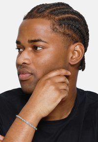 Bracelet argenté torsadé porté au poignet. La personne a les cheveux noirs tressés et porte un t-shirt noir. L'arrière-plan est blanc.
