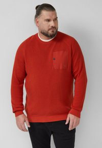 Maglione rosso con texture e design a coste, presenta una tasca sul petto in una tonalità contrastante e un collo tondo. Indossato sopra pantaloni scuri.