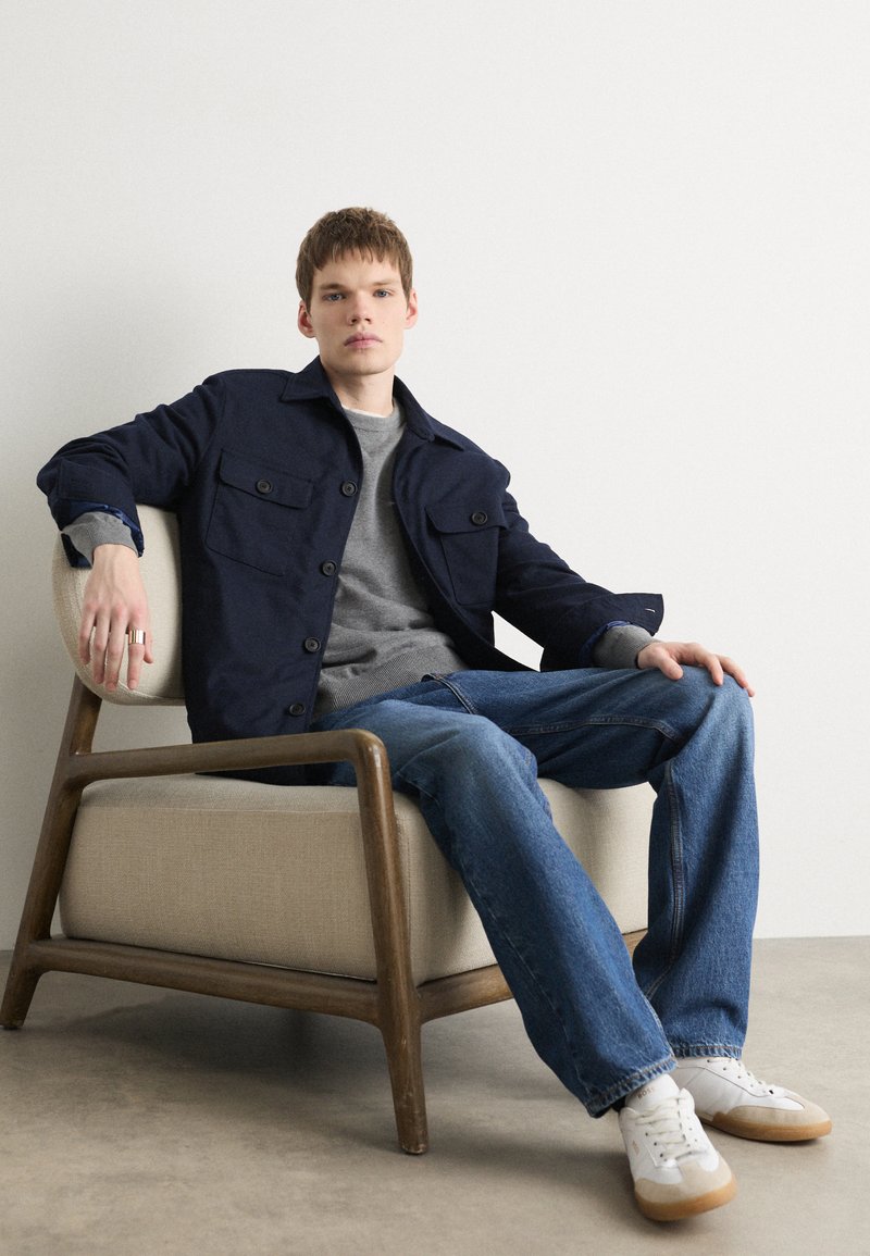 Veste boutonnée bleu marine avec deux poches sur la poitrine, pull gris, jeans bleu clair et baskets blanches avec des accents beige, assis sur une chaise beige.