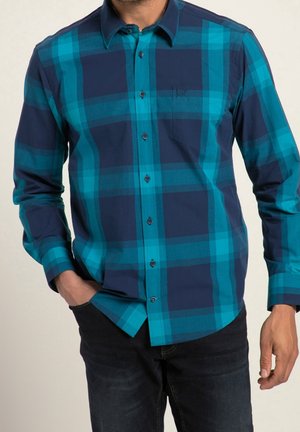 Mann trägt ein kariertes Hemd mit langen Ärmeln in Türkis und Dunkelblau, eine Hand in der Tasche und dunkle Jeans.