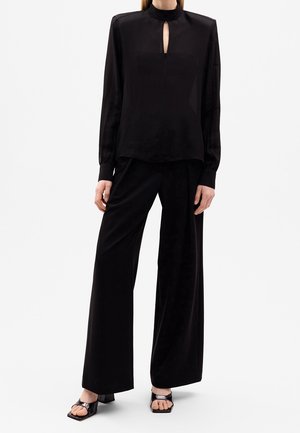 Kobieta nosząca czarną bluzkę z długim rękawem, z wycięciem z przodu, oraz szerokie czarne spodnie, połączone z czarnymi sandałami na obcasie z odkrytymi palcami.