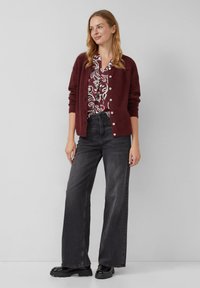 Burgunderfarbener Strickcardigan mit Knöpfen, kombiniert mit einer weiß-burgunderfarbenen Bluse mit Blumenmuster, schwarzen weit geschnittenen Jeans und schwarzen Plattformschuhen.