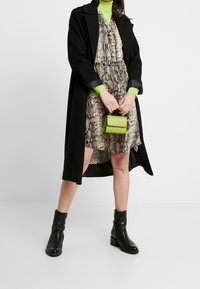 En svart ullkappa över en klänning i ormskinnsmönster, med en grön polotröja och en liten grön handväska. Svarta ankelkängor fulländar outfiten.