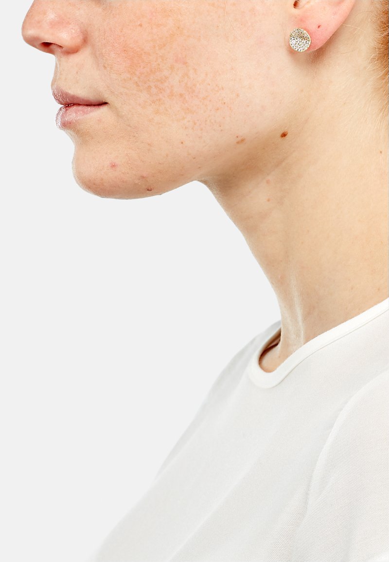 Boucles d'oreilles en argent avec un design circulaire texturé, portées sur un teint de peau clair, avec une chemise blanche unie visible en arrière-plan.