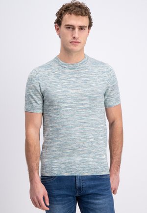 Man met krullend haar, gekleed in een blauw-grijze gebreide trui met korte mouwen en een blauwe spijkerbroek, staand tegen een effen lichte achtergrond.