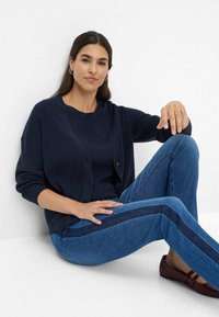Marineblauer,rippierter Cardigan mit Knöpfen, kombiniert mit blauen Jeans mit seitlichem Streifen. Braune flache Schuhe runden das Outfit ab. Einfaches, lässiges Design.