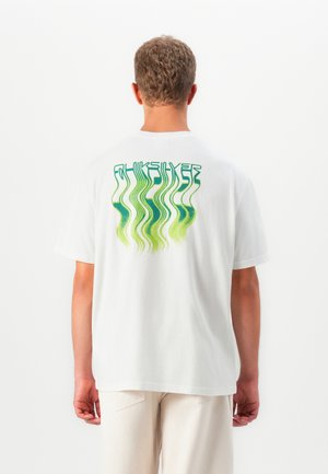 Uomo che indossa una maglietta bianca con un design astratto ondulato verde e del testo sulla schiena, abbinata a pantaloni chiari, in piedi su uno sfondo bianco uniforme.