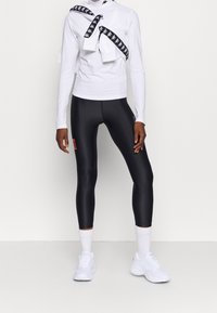 Biała koszula z długim rękawem z czarno-białym paskiem z logo zawiązanym w supeł, w połączeniu z czarnymi legginsami z wysokim stanem i białymi butami sportowymi.