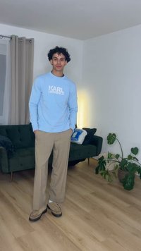 Joven con cabello rizado de pie en el interior, vistiendo una sudadera azul claro, pantalones beige y zapatos beige, junto a un sofá verde y plantas.