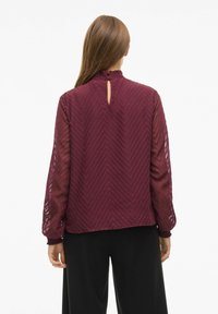 Blusa burdeos con un patrón de chevrón texturizado, detalle de abertura en la espalda y mangas largas transparentes con puños fruncidos, combinada con pantalones negros.
