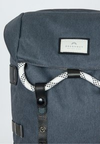 Sac à dos en tissu gris foncé avec fermeture roulée, accents en cuir noir, une sangle tissée blanche et un patch logo sur le devant.