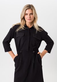 Zwarte shirtjurk met driekwartmouwen, sluiting met knopen aan de voorkant, twee borstzakken en een trekkoord in de taille. Gladde stof met een subtiele glans.