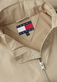 Veste beige avec doublure en mesh, fermeture éclair argentée et étiquette Tommy Jeans indiquant la taille medium à l'intérieur du col.