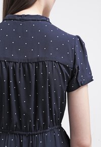 Robe marine à pois blancs, manches courtes bouffantes et taille froncée, avec un détail de bouton sur la manche. Tissu doux.