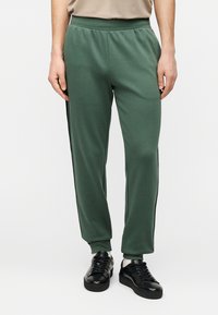 Πράσινο παντελόνι φόρμας με στενή γραμμή, ελαστική μέση και λεπτομέρεια με ρίγα στο πλάι. Κατασκευασμένο από μαλακό, υφασμένο ύφασμα, συνδυασμένο με μαύρα αθλητικά παπούτσια.