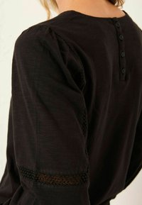 Nahaufnahme einer Person, die ein schwarzes Langarmoberteil mit Rückenschlaufen und Spitze an den Ärmeln und Schultern trägt.