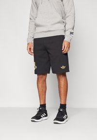 Černé sportovní šortky s detaily zlatého loga, kombinované se šedou mikinou a černými sportovními botami s bílými pruhy. Hladký, ležérní design.