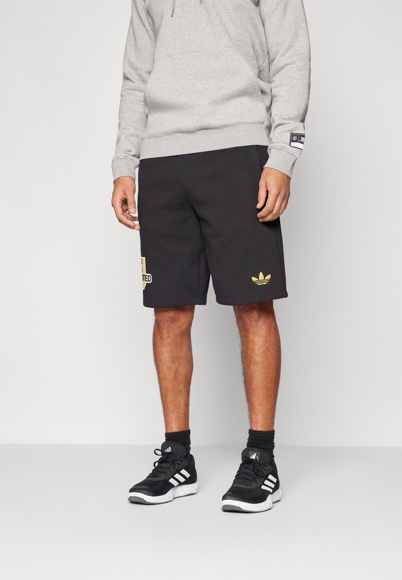 Černé sportovní šortky s detaily zlatého loga, kombinované se šedou mikinou a černými sportovními botami s bílými pruhy. Hladký, ležérní design.