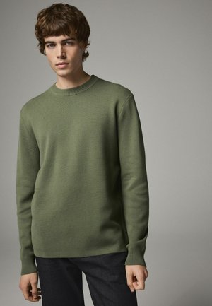 Grøn strikket sweatervare med rund halsudskæring og ribbede manchetter, parret med mørke denimjeans. Enkel design, tekstureret stof og klassisk pasform.