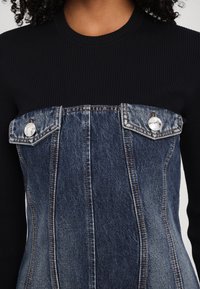 Ribbed černý top s dlouhým rukávem a modrým denimovým panelem, který má stříbrné patentky a strukturovaný design na hrudi.