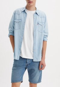 Ljusblå denimskjorta med uppvikta ärmar, två bröstfickor och silverknappar, bärs över en vit t-shirt och kombineras med blå denimshorts.