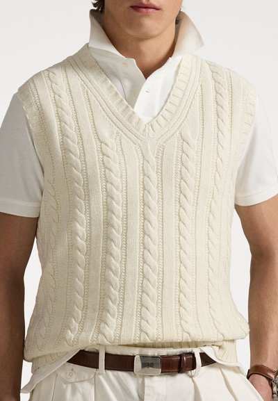 Jersey sin mangas de color crema, tejido a punto con cuello en V, que presenta patrones trenzados texturizados y ribetes acanalados, llevado sobre una camisa blanca con cuello.