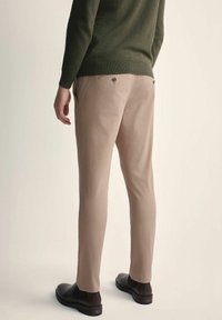 Beżowe spodnie z bawełny, slim fit, o gładkiej teksturze, z dwoma tylnymi kieszeniami zapinanymi na guziki. Noszone z zielonym swetrem.