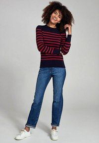 Maglione a righe con base blu navy e righe rosse, abbinato a jeans con risvolto e sneaker bianche. Il maglione ha un collo a giro e polsini a coste.