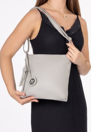 Femme portant une robe noire, tenant un sac à bandoulière en cuir gris clair avec un pompon et un détail d'emblème rond.