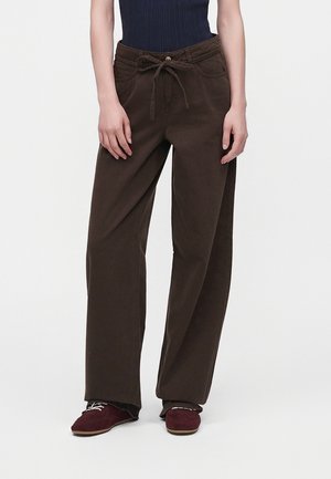Personne portant un pantalon marron taille haute à jambes larges avec une taille à cordon, associé à des chaussures bordeaux lacées et un top sans manches foncé.