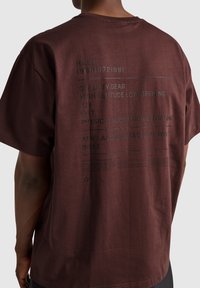 Mörkröd bomullströja med avslappnad passform, korta ärmar och tryckt text på ryggen i svart. Designen inkluderar olika militära termer.