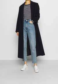 Cappotto lungo di lana blu navy, maglia a righe, jeans a sigaretta azzurri chiari e sneakers bianche. Il cappotto presenta un colletto a risvolto e dettagli con bottoni.
