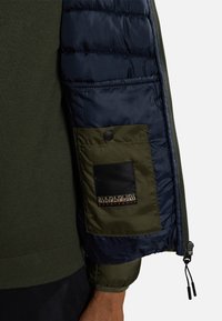 Giacca imbottita con un guscio in nylon blu scuro e polsini in maglina verde oliva. Presenta una tasca a toppa con bottone e un dettaglio del logo sulla manica.