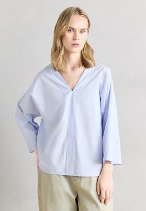 Mujer rubia con blusa de manga larga de cuello en V azul claro extragrande y pantalones beige, sobre un fondo liso gris claro.