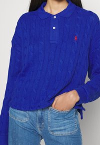 Blå kabelstickad polo skjorta med krage och treknapps placket. Har en liten röd logga på bröstet och ribbade ärmslut. Kombinerad med blå jeans.