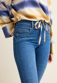Jean en denim bleu avec une coupe ajustée, présentant une poche avant, des détails de couture subtils et une texture lisse. Assorti à un blouse à motif.