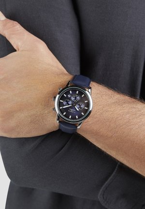 Man met zwarte mouw toont polshorloge met marineblauwe band en zwarte wijzerplaat met drie kleinere chronograaf-wijzerplaten en een datumvenster.