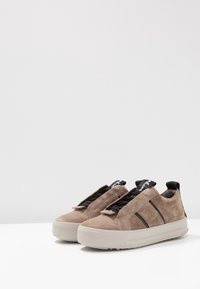 Beige suede tennarit, joissa on paksu valkoinen pohja, mustia korostusraitaja ja joustavat nauhat. Muotoilussa on myös vetoketju takana.