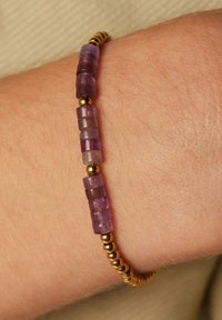 Des perles de verre violet en forme de cube, entrecoupées de perles séparatrices dorées, forment un bracelet reposant sur une peau de ton clair. Texture lisse et forme arrondie.