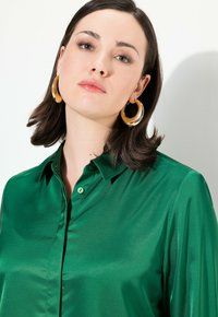 Zielona satynowa koszula z długim rękawem, z kołnierzykiem, gładką fakturą i pojedynczym guzikiem przy kołnierzyku.